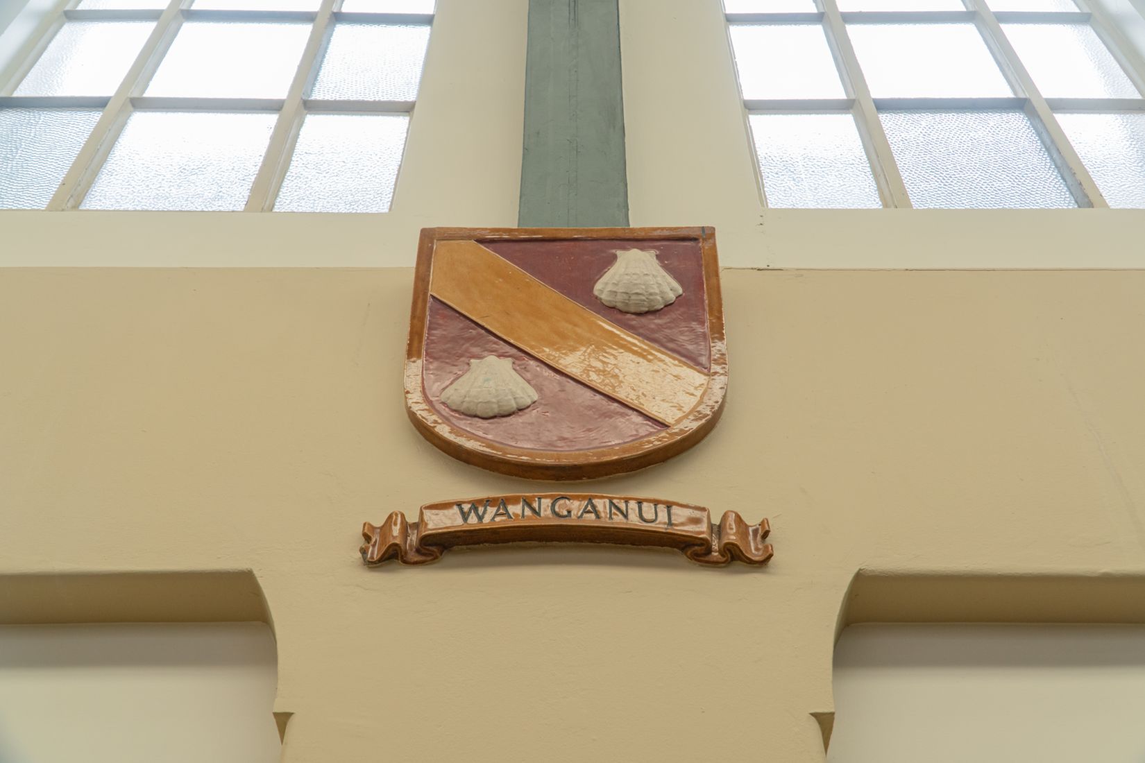 A painted Wanganui provincial coat of arms mounted on the hall wall beneath a heritage window.