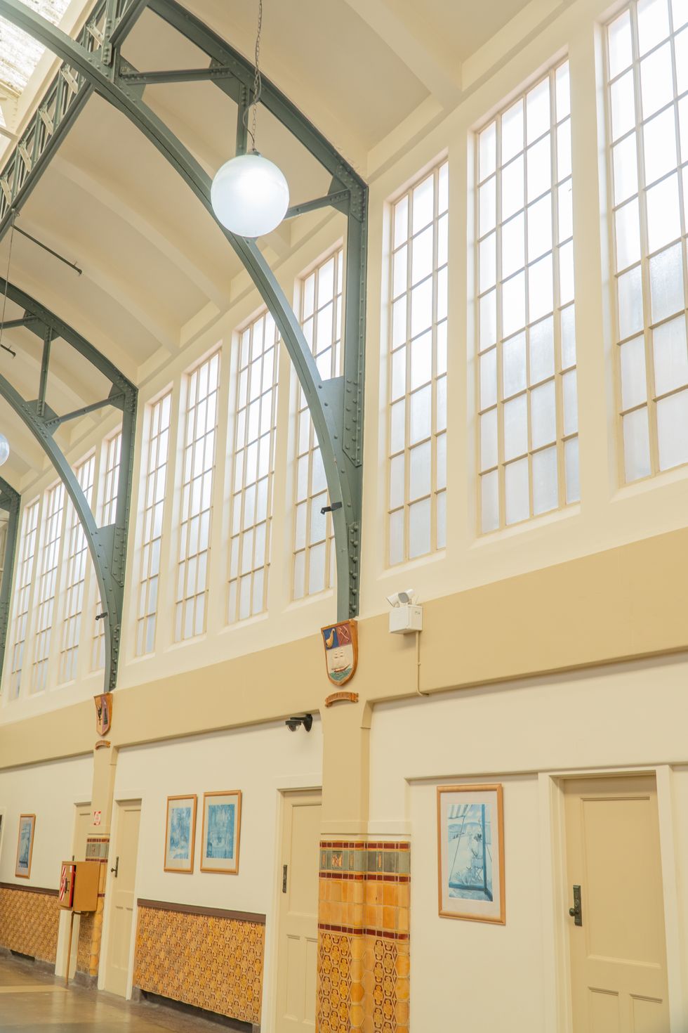 A riveted steel arch and globe pendant beside the hall's tall multi-paned windows and provincial crests.
