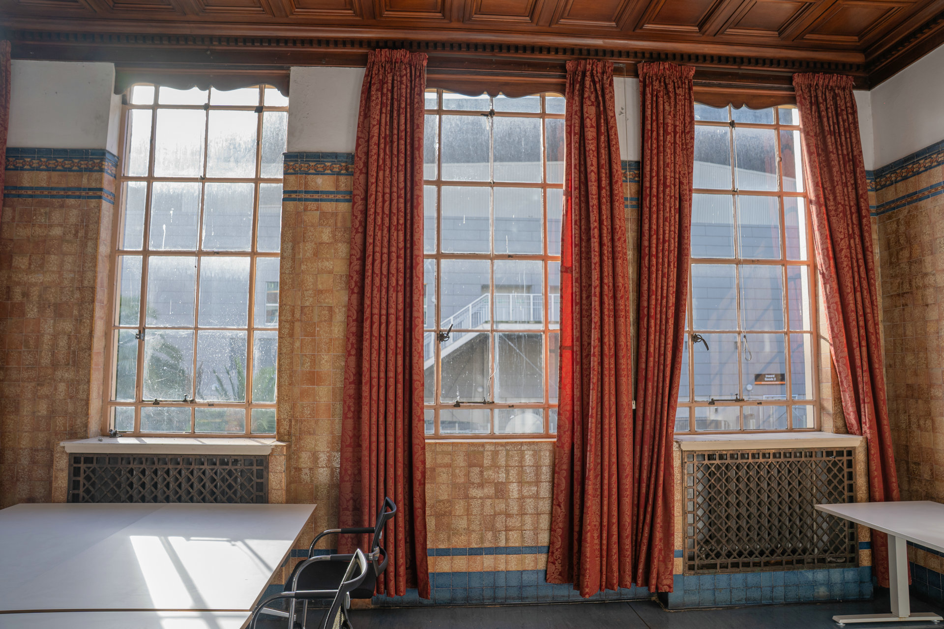 Tall timber-framed windows with red damask curtains above glazed tiles and cast-iron radiator covers.